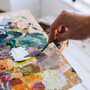 May include: A wooden artist's palette with a variety of mixed paint colors, including white, green, brown, and yellow. A hand holds a metal palette knife, spreading white paint. The palette is on a wooden easel.