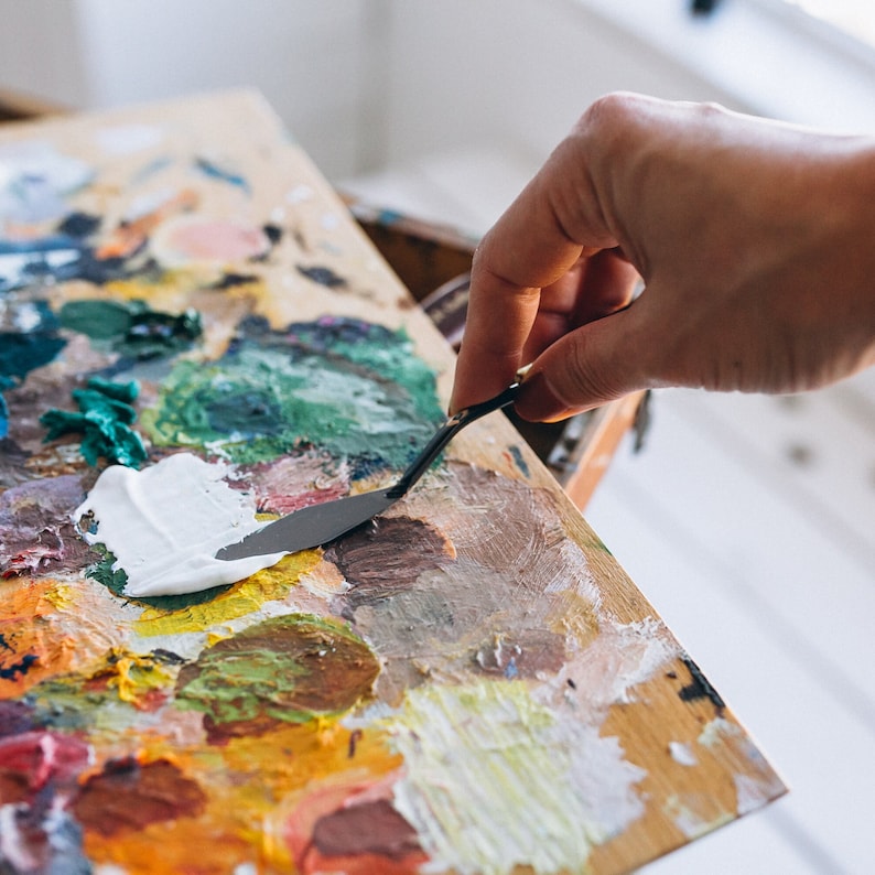 Puede incluir: Una mano sosteniendo una esp&aacute;tula de paleta de metal, raspando pintura blanca de una paleta de madera con varios colores de pintura.