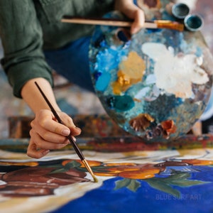 May include: An artist's hands holding a paintbrush and a wooden palette filled with various colors of paint. The artist is working on a painting with blue and brown tones. The painting has the text "BLUE SURF ART" in the bottom right corner.