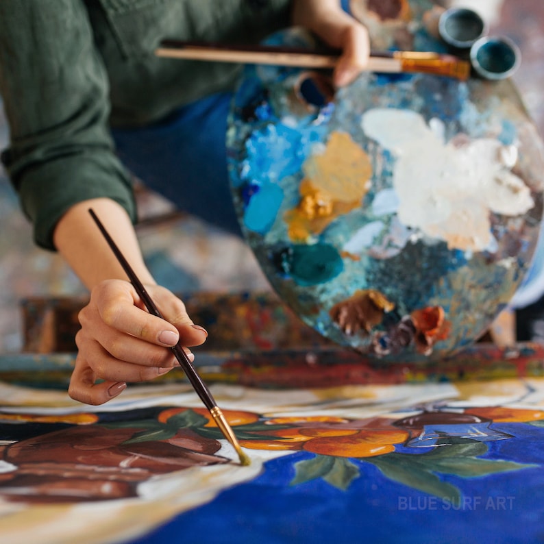 May include: An artist's hands holding a paintbrush and a wooden palette filled with various colors of paint. The artist is working on a painting with blue and brown tones. The painting has the text "BLUE SURF ART" in the bottom right corner.