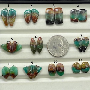 May include: A collection of eleven polished gemstones in a black tray. The stones display a variety of shapes and colors, including teal, brown, and green. A US quarter is included for size reference. The gemstones are numbered 1 through 11.