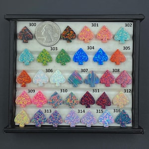 Può includere: Una collezione di cabochon a forma di picche in vari colori e finiture iridescenti, disposti in un vassoio espositore nero. Una moneta da 25 centesimi americana è inclusa per la scala. Ogni pezzo è etichettato con un numero da 300 a 316.