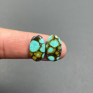 May include: Two oval-shaped turquoise gemstones with a matrix of brown and gold. The gemstones are held between fingers against a gray background. The turquoise stones have a unique pattern and are polished.