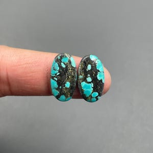 May include: Two oval-shaped turquoise gemstones with a matrix of black and brown. The gemstones have a polished surface and are held between fingers against a gray background. The turquoise has a speckled pattern.