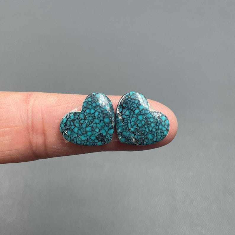 May include: Two heart-shaped turquoise stones with a mosaic pattern of dark blue and black veins. The stones are held between two fingers against a gray background. The turquoise has a polished, glossy finish.