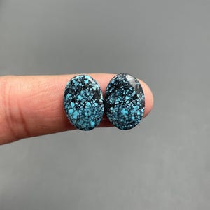 May include: Two oval-shaped turquoise gemstones with a black matrix. The gemstones are held between fingers against a gray background. The turquoise has a speckled appearance, with varying shades of blue and black.
