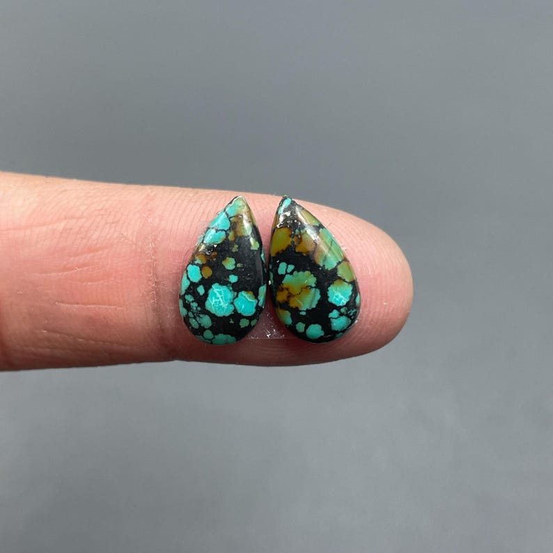 May include: Two teardrop-shaped turquoise gemstones with a black matrix and brown accents. The gemstones have a polished surface and are displayed against a gray background. The turquoise stones are likely for jewelry making.