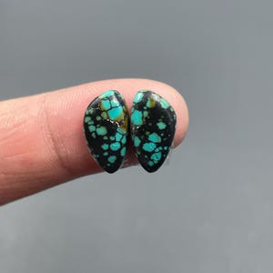 May include: Two teardrop-shaped turquoise gemstones with a black matrix. The gemstones have a speckled pattern of bright turquoise against a dark background. The stones are displayed against a neutral gray backdrop.