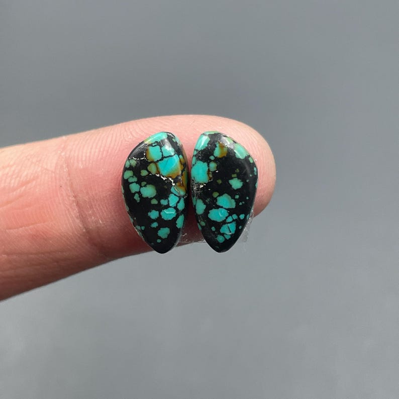May include: Two teardrop-shaped turquoise gemstones with a black matrix. The gemstones have a speckled pattern of bright turquoise against a dark background. The stones are displayed against a neutral gray backdrop.