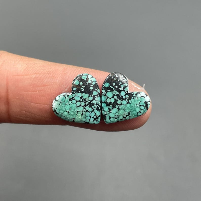 May include: Two heart-shaped turquoise gemstones with a speckled pattern of teal and black. The gemstones are held between fingers, showcasing their unique color and texture. These could be used for jewelry making.