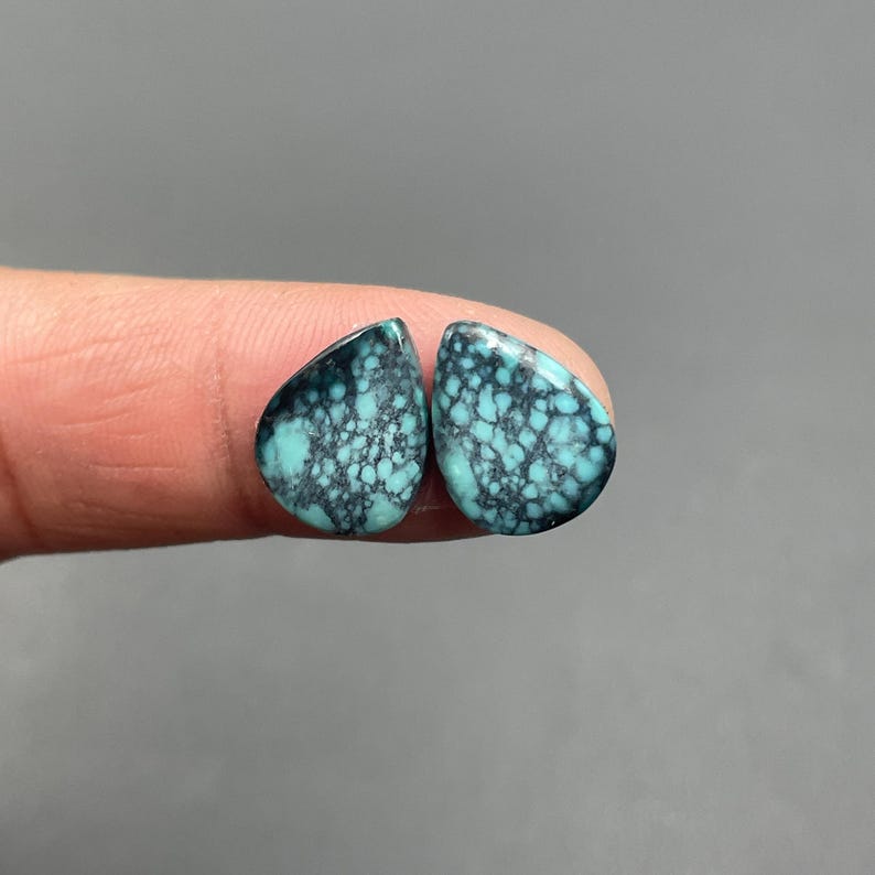 May include: Two teardrop-shaped turquoise gemstones with a dark matrix pattern. The stones are a vibrant teal color with a network of dark gray veins. The gemstones are displayed against a neutral gray background.
