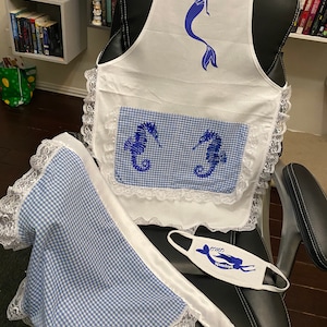 May include: A white apron with blue embroidered seahorses and a mermaid design. The apron has a gingham pocket and lace trim. A matching blue and white gingham fabric with lace trim is also pictured. A white face mask with a blue mermaid design and the text "omo remaya" is also pictured.