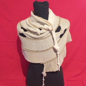 May include: Cream-colored knitted shawl with a unique design. The shawl features decorative knots and horizontal lines with a contrasting beige trim. The garment is displayed on a black mannequin against a red background. A stylish accessory for any wardrobe.