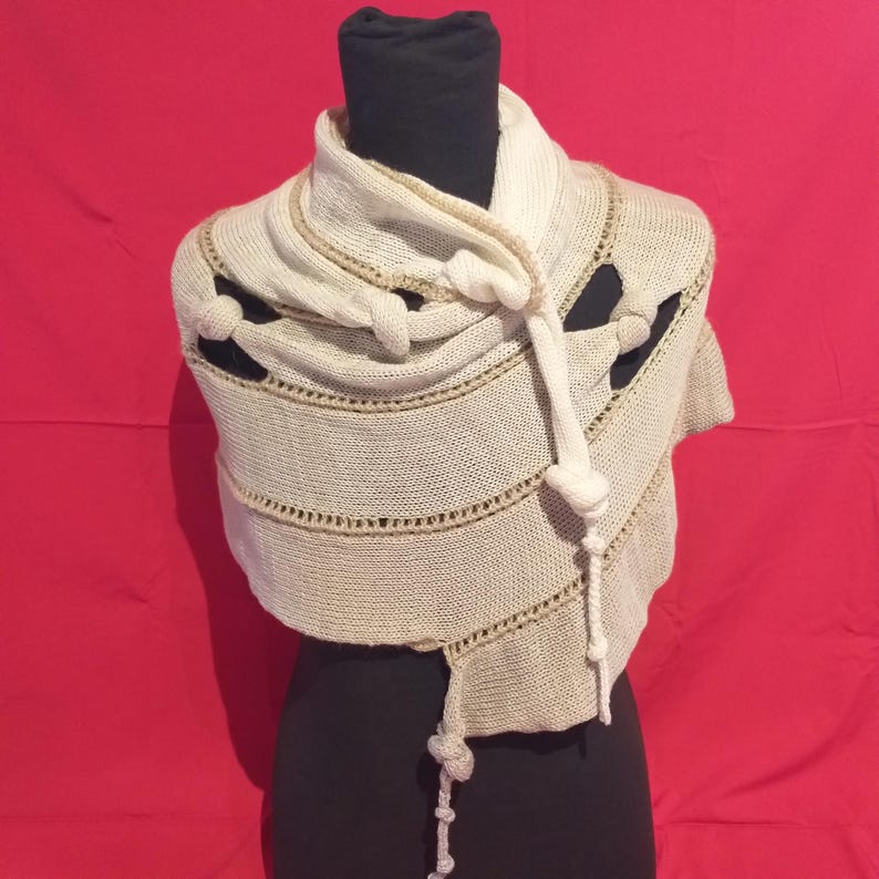 May include: Cream-colored knitted shawl with a unique design. The shawl features decorative knots and horizontal lines with a contrasting beige trim. The garment is displayed on a black mannequin against a red background. A stylish accessory for any wardrobe.