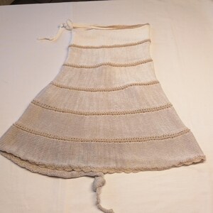 May include: A cream-colored, knitted skirt with horizontal tan stripes. The skirt has a flared shape and a scalloped hemline. A drawstring is visible at the top and a knotted tie at the bottom. The skirt appears to be handmade.