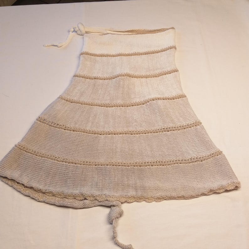 May include: A cream-colored, knitted skirt with horizontal tan stripes. The skirt has a flared shape and a scalloped hemline. A drawstring is visible at the top and a knotted tie at the bottom. The skirt appears to be handmade.