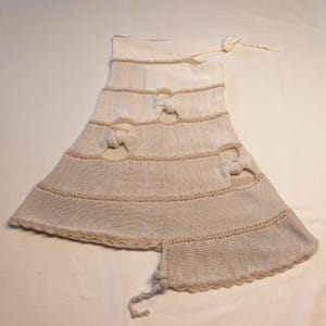 May include: A cream-colored, knitted garment with a flared skirt and decorative bow accents. The piece features horizontal bands with knotted details and a scalloped edge. A long, knotted tie extends from the top.