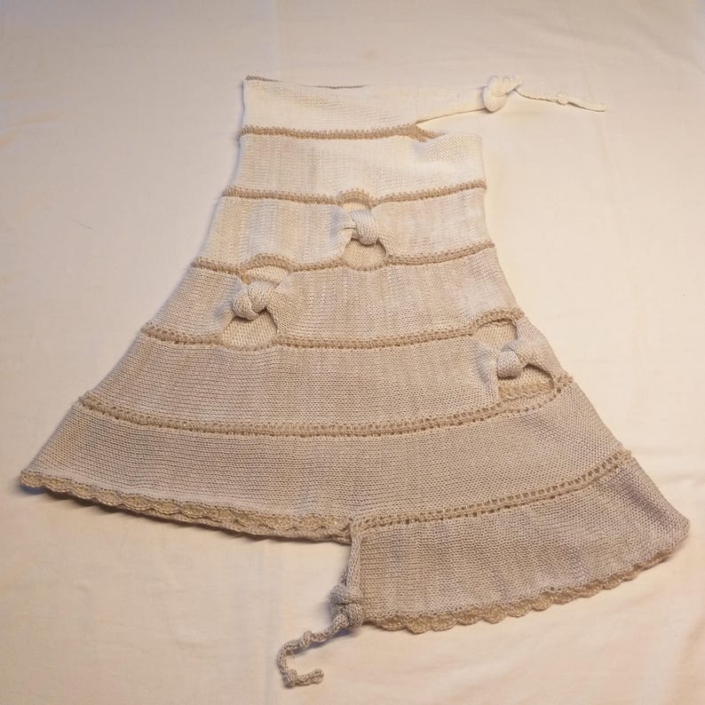 May include: A cream-colored, knitted garment with a flared skirt and decorative bow accents. The piece features horizontal bands with knotted details and a scalloped edge. A long, knotted tie extends from the top.