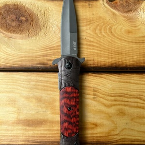 Puede incluir: Un cuchillo plegable negro con mango de madera rojo y negro. La hoja es de estilo daga y el mango tiene la palabra "JEEP" y "DA145" grabadas.