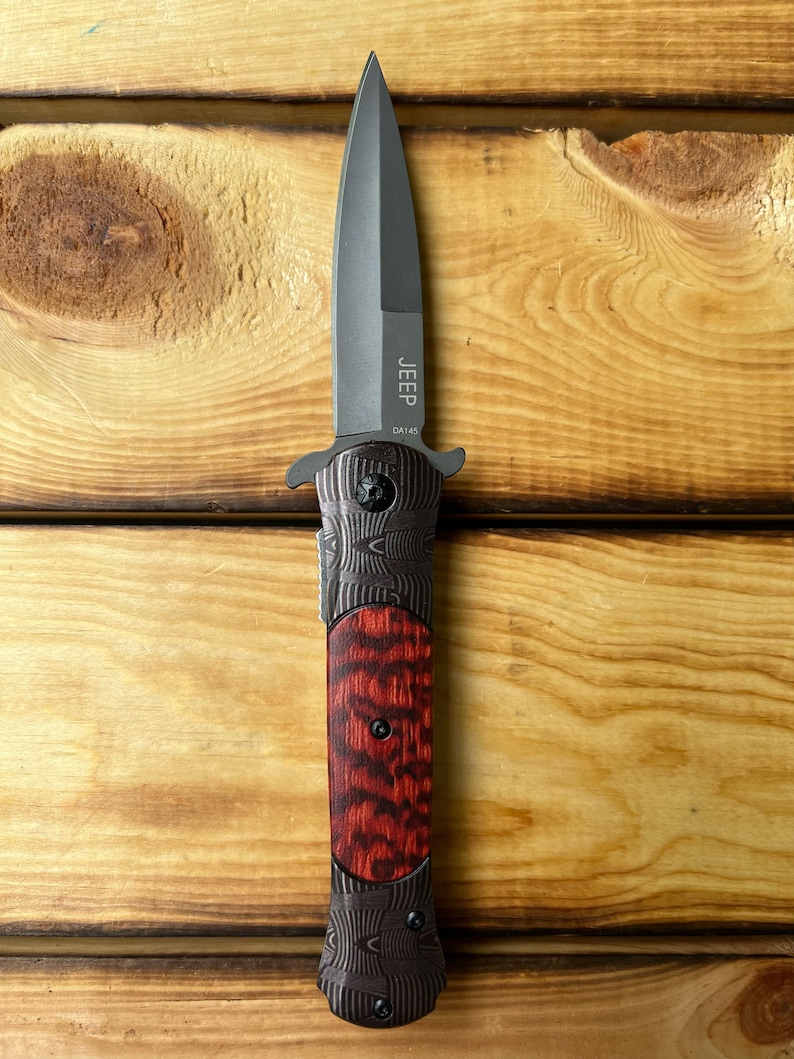 Puede incluir: Un cuchillo plegable negro con mango de madera rojo y negro. La hoja es de estilo daga y el mango tiene la palabra "JEEP" y "DA145" grabadas.