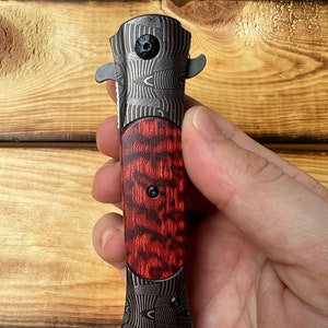 Puede incluir: Un cuchillo de bolsillo negro y rojo con un mango estampado. El mango tiene un patr&oacute;n de veta de madera con acentos rojos y negros. La hoja est&aacute; cerrada y el cuchillo se sostiene en una mano.