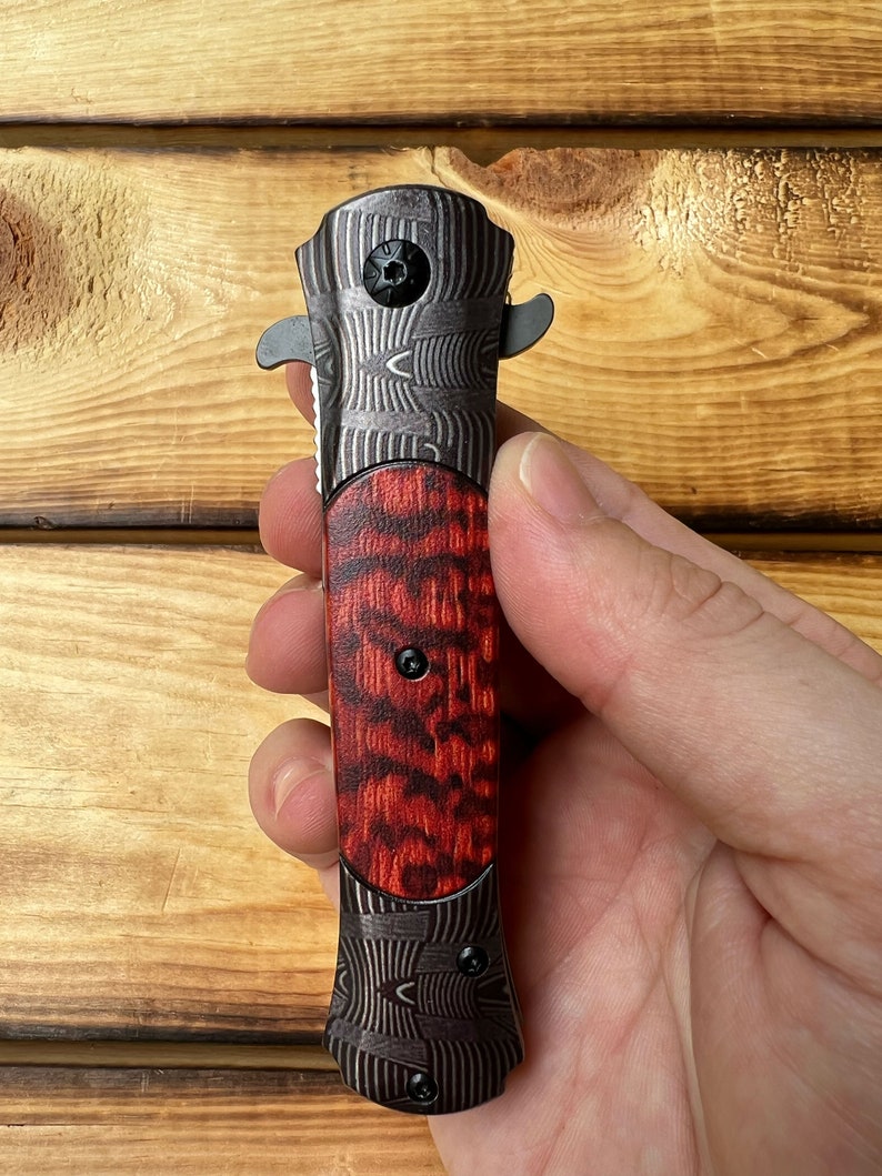 Puede incluir: Un cuchillo de bolsillo negro y rojo con un mango estampado. El mango tiene un patr&oacute;n de veta de madera con acentos rojos y negros. La hoja est&aacute; cerrada y el cuchillo se sostiene en una mano.