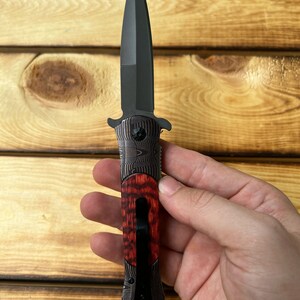 Puede incluir: Un cuchillo plegable negro con mango de madera rojo y negro. La hoja es de estilo daga de doble filo.