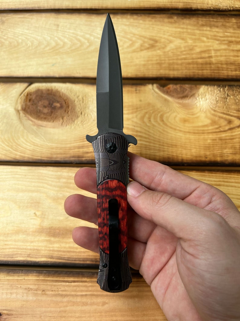 Puede incluir: Un cuchillo plegable negro con mango de madera rojo y negro. La hoja es de estilo daga de doble filo.