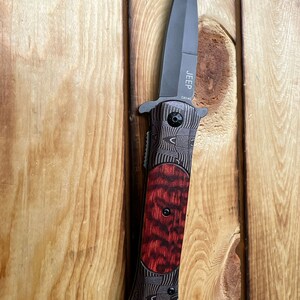 Puede incluir: Un cuchillo plegable negro con un mango de madera rojo y negro. La hoja est&aacute; parcialmente abierta y el cuchillo est&aacute; sujeto a una superficie de madera. El cuchillo tiene la palabra "JEEP" y las letras "DA145" grabadas en la hoja.