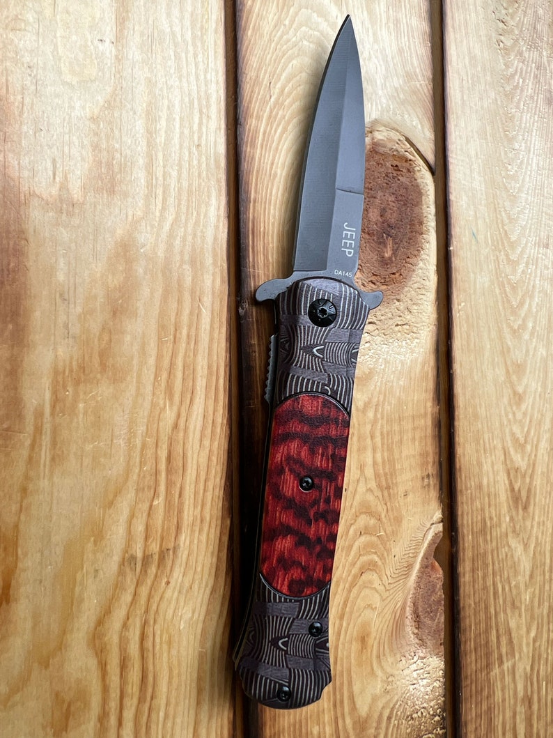 Puede incluir: Un cuchillo plegable negro con un mango de madera rojo y negro. La hoja est&aacute; parcialmente abierta y el cuchillo est&aacute; sujeto a una superficie de madera. El cuchillo tiene la palabra "JEEP" y las letras "DA145" grabadas en la hoja.
