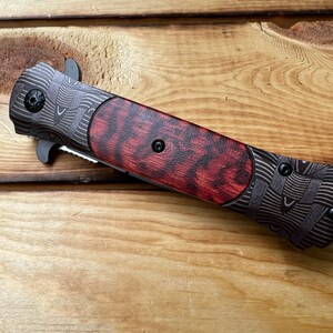 Puede incluir: Un cuchillo de bolsillo negro y rojo con un mango estampado. El mango es de madera y tiene un patr&oacute;n rojo y negro. La hoja est&aacute; cerrada y el cuchillo est&aacute; sobre una superficie de madera.