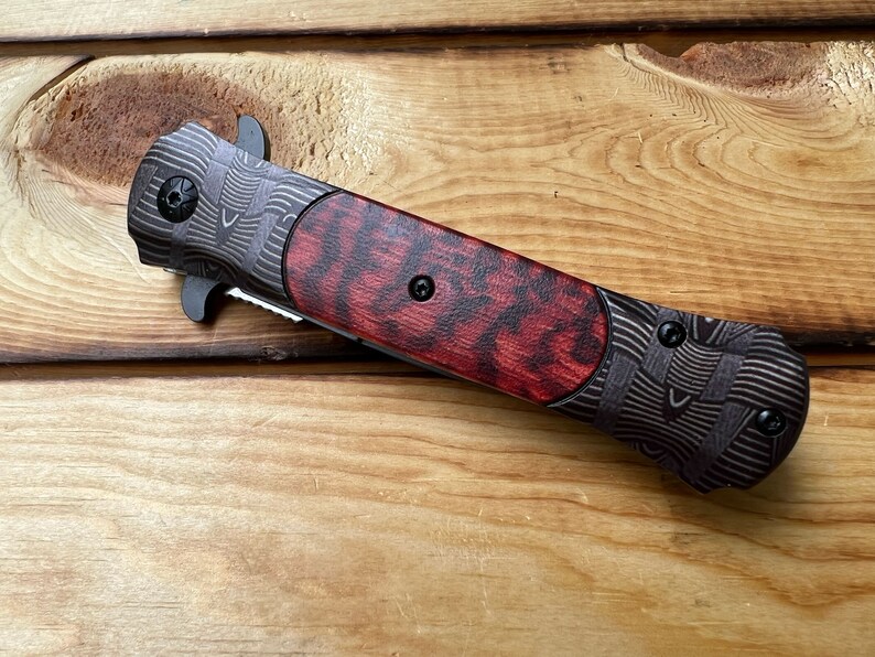 Puede incluir: Un cuchillo de bolsillo negro y rojo con un mango estampado. El mango es de madera y tiene un patr&oacute;n rojo y negro. La hoja est&aacute; cerrada y el cuchillo est&aacute; sobre una superficie de madera.