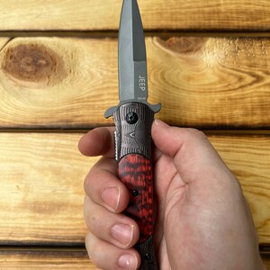 Puede incluir: Un cuchillo de bolsillo negro y rojo con una hoja negra y un mango con un patr&oacute;n rojo y negro. El cuchillo tiene la palabra "JEEP" y "DA145" grabadas en la hoja.