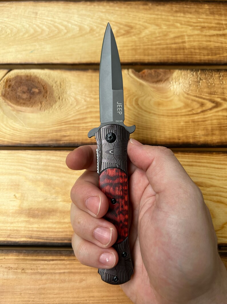 Puede incluir: Un cuchillo de bolsillo negro y rojo con una hoja negra y un mango con un patr&oacute;n rojo y negro. El cuchillo tiene la palabra "JEEP" y "DA145" grabadas en la hoja.