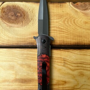 Puede incluir: Un cuchillo plegable negro con mango de madera rojo y negro. La hoja es de estilo daga de doble filo.