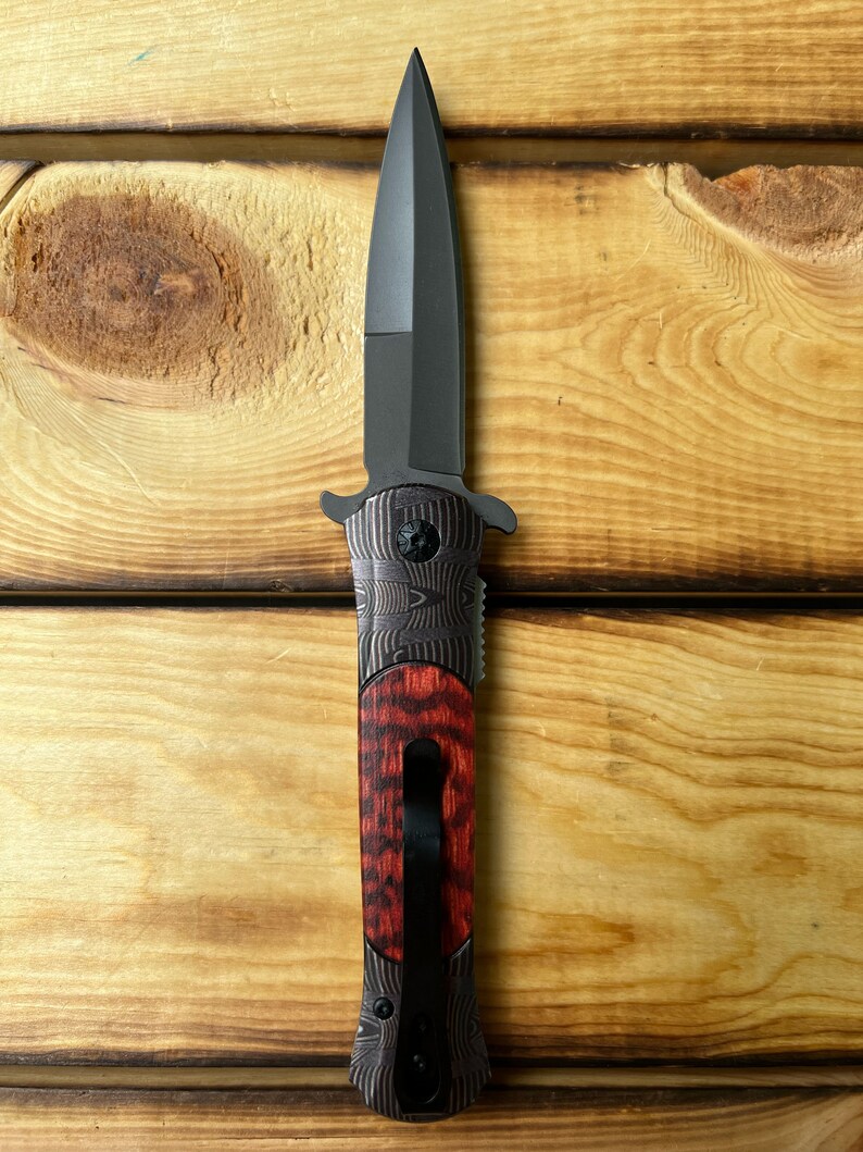 Puede incluir: Un cuchillo plegable negro con mango de madera rojo y negro. La hoja es de estilo daga de doble filo.