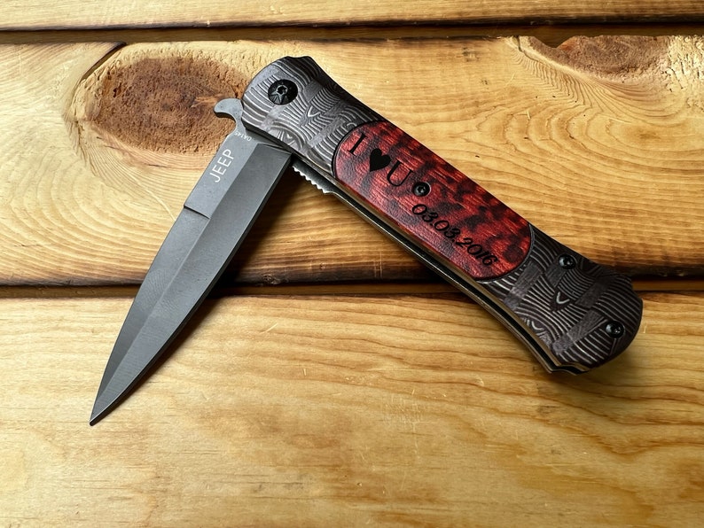 Puede incluir: Un cuchillo de bolsillo negro y gris con un mango de madera rojo y negro. El mango est&aacute; grabado con "I love you 08.03.2016" y la hoja est&aacute; grabada con "JEEP".