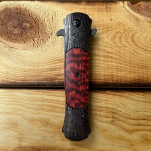 Puede incluir: Un cuchillo de bolsillo negro y rojo con un mango estampado. El mango est&aacute; hecho de madera y metal.
