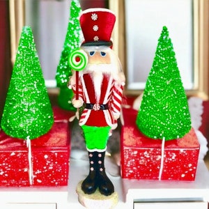May include: A red and white striped nutcracker figurine holding a green and white lollipop. The nutcracker is standing in front of two green, sparkly Christmas trees.