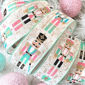 May include: Cream ribbon featuring a repeating pattern of nutcracker soldiers in pink, teal, and gold. The ribbon has a mint green border and gold swirl accents, ideal for festive crafts and decorations.