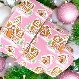 May include: Pink ribbon with a repeating pattern of gingerbread houses, candy canes, and green trees. The ribbon is rolled up and ready to be used for wrapping gifts.