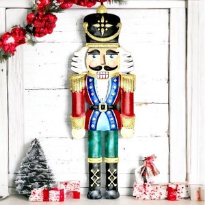 May include: A decorative wooden nutcracker soldier in a festive Christmas display. The nutcracker is painted in red, blue, gold, and green, with a black hat and white hair. The display includes a small Christmas tree and wrapped gifts.
