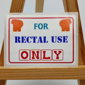 Peut inclure: Un autocollant blanc avec du texte rouge et bleu qui dit "FOR RECTAL USE ONLY". L'autocollant est sur un petit chevalet en bois.