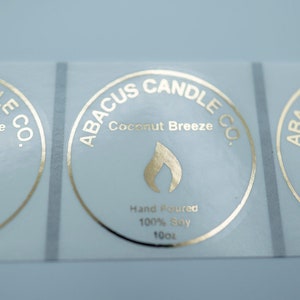 May include: Three gold foil labels with the text "ABACUS CANDLE CO." and "Coconut Breeze" in a circle. The labels also include the text "Hand Poured 100% Soy 10oz".