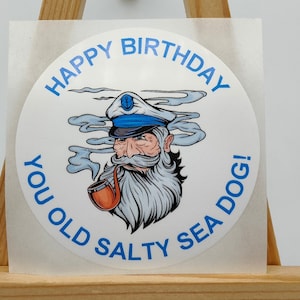 Peut inclure: Un autocollant rond blanc avec une bordure bleue et le texte "Happy Birthday You Old Salty Sea Dog!" en bleu. Une illustration de dessin animé d'un marin avec une pipe et une barbe blanche est au centre de l'autocollant.