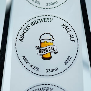 May include: A roll of white round stickers with a black and white design. The stickers feature a beer glass with the text "Beer Day" and the words "Abacus Brewery Pale Ale" around the glass. The stickers also include the text "ABV: 4.5% 330ml 2022".