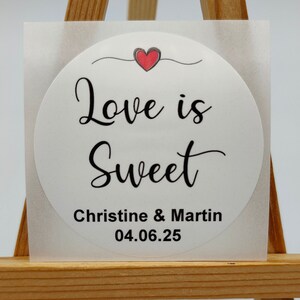 May include: White round sticker with the text "Love is Sweet" in black cursive font. A small red heart is above the text. Below the text is "Christine & Martin" and "04.06.25" in black font.