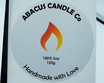 May include: Two white round labels with black text and a flame graphic. The top label reads "100% Soy 120g Handmade with Love". The bottom label reads "ABACUS CANDLE Co 100% Soy 120g Handmade with Love".