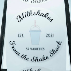 May include: White round sticker with black text that reads "Milkshakes from the Shake Shack" with a stylised image of a milkshake cup with a straw. The text also includes "EST. 2021" and "57 Varieties".