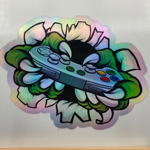 May include: A holographic sticker of a green monster hand breaking through a wall, holding a video game controller.